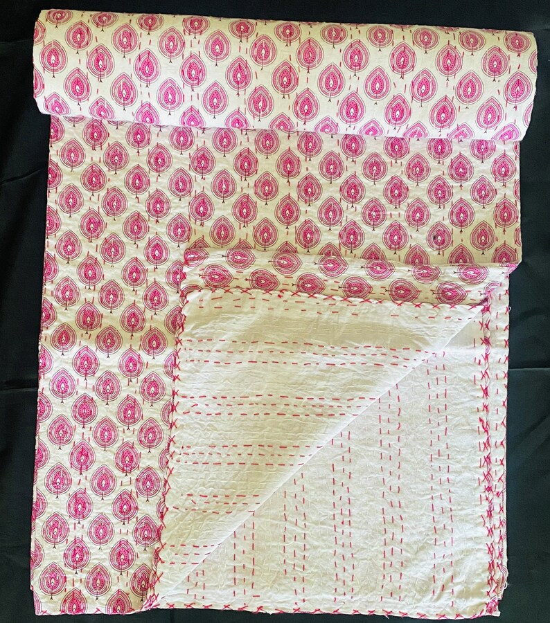 May include: A rolled-up, patterned textile with a white background and a repeating pink leaf design. The unfolded portion reveals a white, quilted fabric with red stitching. The textile is likely a blanket or throw.