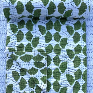 May include: A white kantha quilt with a green ginkgo leaf pattern. The quilt is folded in half and is lying on a blue and white patterned surface.