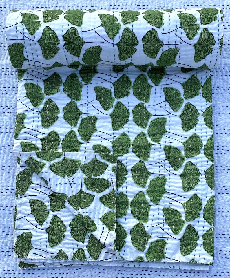 May include: A white kantha quilt with a green ginkgo leaf pattern. The quilt is folded in half and is lying on a blue and white patterned surface.