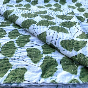 Può includere: Una trapunta in cotone bianco con un motivo ripetuto di foglie di ginkgo verdi con contorni neri. Il tessuto è rifinito con visibili impunture orizzontali e verticali in filo verde e bianco, che creano un aspetto trapuntato e testurizzato.