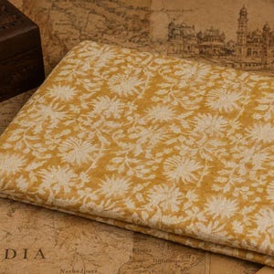 May include: Folded yellow fabric with a white floral design. The fabric is neatly folded and appears to be made of a soft material. The background includes a wooden box and a map of India.