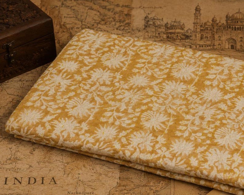 May include: Folded yellow fabric with a white floral design. The fabric is neatly folded and appears to be made of a soft material. The background includes a wooden box and a map of India.