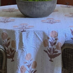 May include: A white tablecloth with a repeating floral pattern in shades of brown and peach. The flowers have a stylized design. The tablecloth is draped over a round table, with a gray bowl filled with greenery in the background.