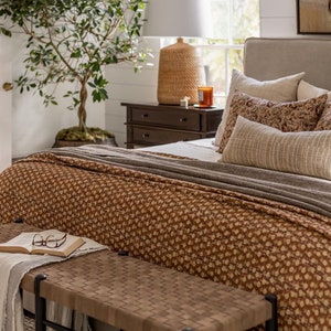 Op de afbeelding: Een bruine houten bank met een geweven leren bovenkant staat voor een bed met een bruin en wit gestreept dekbedovertrek. Het bed heeft een beige hoofdeinde en verschillende kussens. Een klein houten nachtkastje met drie laden staat naast het bed, met een lamp en een kaars erop.