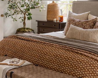May include: A brown wooden bench with a woven leather top sits in front of a bed with a brown and white patterned duvet cover. The bed has a beige headboard and several pillows. A small wooden nightstand with three drawers is next to the bed, with a lamp and a candle on top.