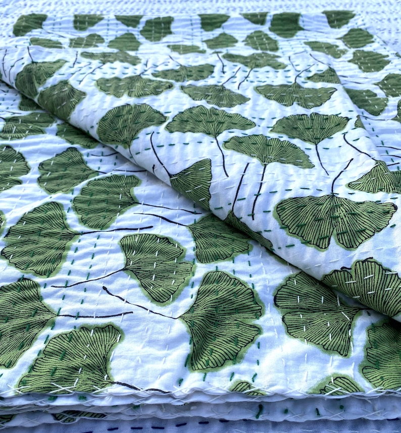 May include: White cotton kantha quilt with a green ginkgo leaf pattern. The quilt is folded in half and the leaves are stitched in a repeating pattern.