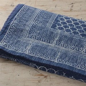 May include: A folded rectangular textile featuring a deep blue base with white block-printed patterns. The design includes a scalloped border, a wavy line motif, and a repeating fish-scale pattern, all finished with visible white running stitch quilting.