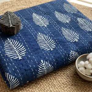 May include: A folded dark blue quilt featuring a white fern leaf pattern and white running stitch details. A carved wooden block with a matching leaf design rests on the fabric, and a small bowl contains raw cotton bolls and a metal thimble.