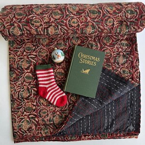 May include: A folded dark red quilt with a repeating pomegranate pattern and white kantha stitching, featuring a green book titled Christmas Stories, a small round ornament with a deer illustration, and a red and white striped knit stocking.