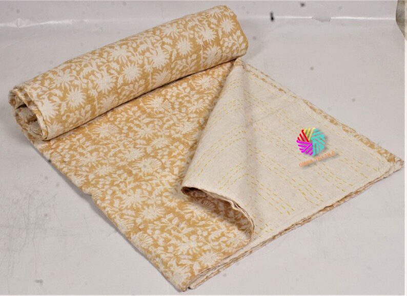May include: A rolled-up, mustard-yellow and cream-coloured quilt with a floral pattern. The quilt is partially unfolded, revealing a cream-coloured underside with stitched lines. The quilt is made of soft fabric.