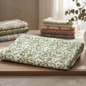 K&ouml;nnte beinhalten: Ein gefaltetes wei&szlig;es Textil mit einem gr&uuml;nen Blumenmuster, m&ouml;glicherweise eine Tischdecke oder ein Platzset, liegt auf einer Holzoberfl&auml;che. Weitere gefaltete Textilien mit &auml;hnlichen Mustern in verschiedenen Farben sind im Hintergrund gestapelt. Das Design zeigt gr&uuml;ne Ranken und florale Elemente.