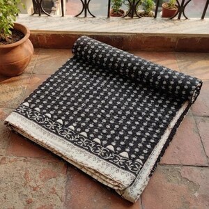 May include: A folded black and white cotton quilt featuring a repeating floral block print pattern. The textile displays a dense arrangement of small flowers across the main surface, bordered by a wider band of intricate botanical motifs.