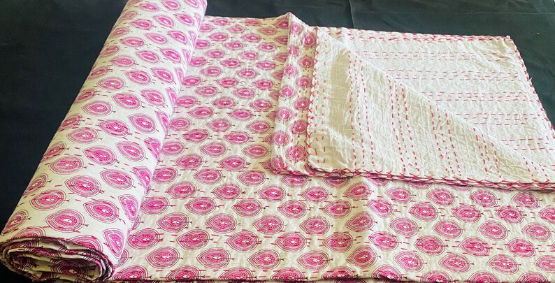 May include: A rolled-up white and pink quilt with a repeating leaf pattern. The quilt is hand-stitched with pink thread, and a portion is unfolded, revealing the reverse side with vertical stitching. The overall theme is home decor.