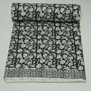 May include: A rolled-up black and white floral printed fabric. The fabric is covered in a repeating pattern of white flowers and leaves on a black background. The edge of the fabric has a decorative border.
