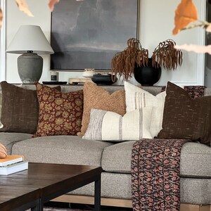 May include: A gray couch with a variety of throw pillows, including a brown patterned pillow, a tan pillow, a white pillow with black stripes, and a brown pillow with a woven texture. A black and white patterned throw blanket is draped over the couch. A wooden coffee table with a black metal frame sits in front of the couch.