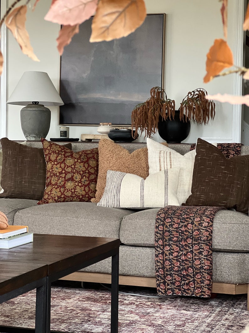May include: A gray couch with a variety of throw pillows, including a brown patterned pillow, a tan pillow, a white pillow with black stripes, and a brown pillow with a woven texture. A black and white patterned throw blanket is draped over the couch. A wooden coffee table with a black metal frame sits in front of the couch.