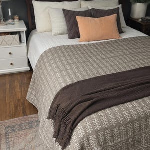 May include: A bed featuring a taupe quilt with a white repeating botanical pattern, topped with a dark brown fringed throw blanket. The bedding includes two white pillows, two dark grey pillows, and one rectangular peach-coloured accent pillow.