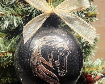 May include: A black Christmas ornament with a gold horse head design and a gold glitter bow. The ornament is hanging on a Christmas tree with green needles.