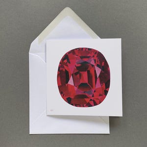 May include: A greeting card featuring a detailed illustration of a faceted, oval-shaped ruby in shades of red and pink. The card is inside a white envelope, set against a gray background. The artist's initials, "AJG", are in the lower left corner.