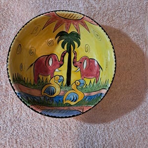 May include: A hand-painted ceramic bowl with a vibrant African savanna scene. The bowl features two pink elephants, a palm tree, and two yellow birds. The background is yellow with a sun design. The rim is black.