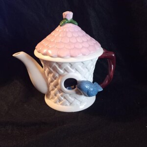 May include: A white ceramic teapot shaped like a birdhouse with a pink roof and a blue bird perched on the side. The teapot has a brown handle and a small hole in the front.