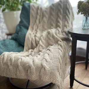 May include: A cream-colored knitted throw blanket with a cable knit pattern draped over a teal armchair. The blanket is soft and cozy, perfect for adding warmth and style to any room.