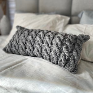 Può includere: Un cuscino grigio lavorato a maglia con un motivo a treccia. Il cuscino è appoggiato su un letto bianco con un motivo geometrico.