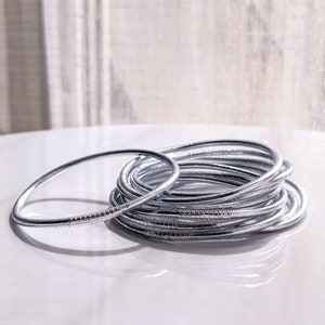 Puede incluir: Una pila de pulseras plateadas, algunas con una superficie texturizada, sobre una superficie blanca.