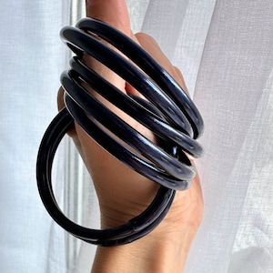 Könnte beinhalten: Ein Set aus fünf schwarzen Armreifen, die auf dem Handgelenk einer Person übereinander liegen. Die Armreifen sind aus einem glatten, glänzenden Material gefertigt.