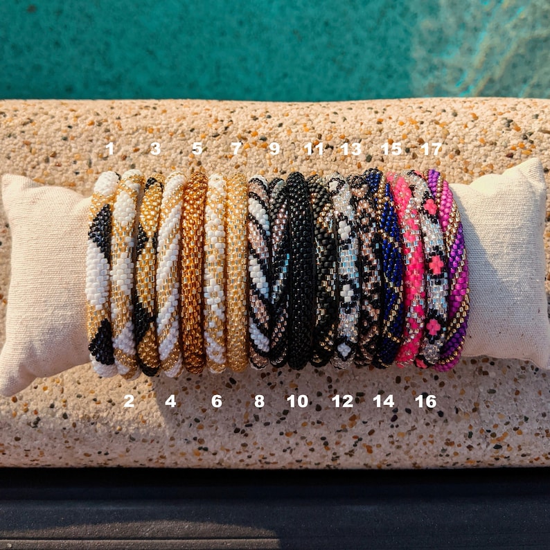 May include: A collection of 17 colourful beaded bracelets, stacked on a white fabric surface. The bracelets are made with various patterns and colours, including black, white, gold, pink, and blue.
