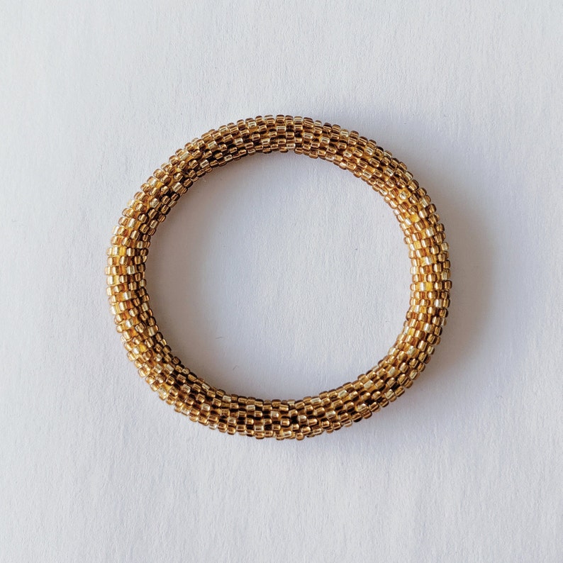 May include: A gold beaded bracelet with a round shape. The bracelet is made of small, square beads that are woven together.