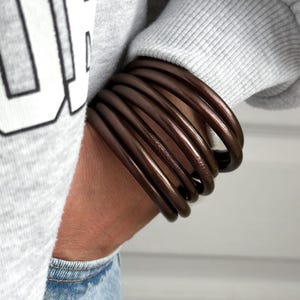 Può includere: Un set di braccialetti rigidi marroni realizzati in un materiale lucido e flessibile. I braccialetti sono indossati al polso.