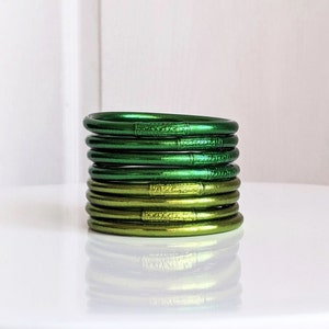 Op de afbeelding: Een stapel groene en gouden bangle armbanden. De armbanden zijn gemaakt van een glanzend, metalen materiaal en hebben een subtiele textuur.