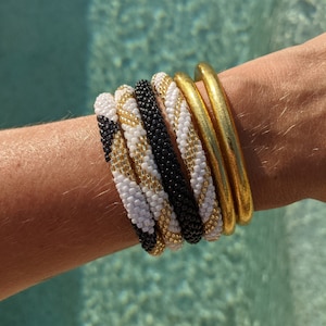 May include: A stack of colourful bracelets, including black, white, and gold, worn on a person's wrist. The bracelets are made of small beads and are stacked on top of each other.