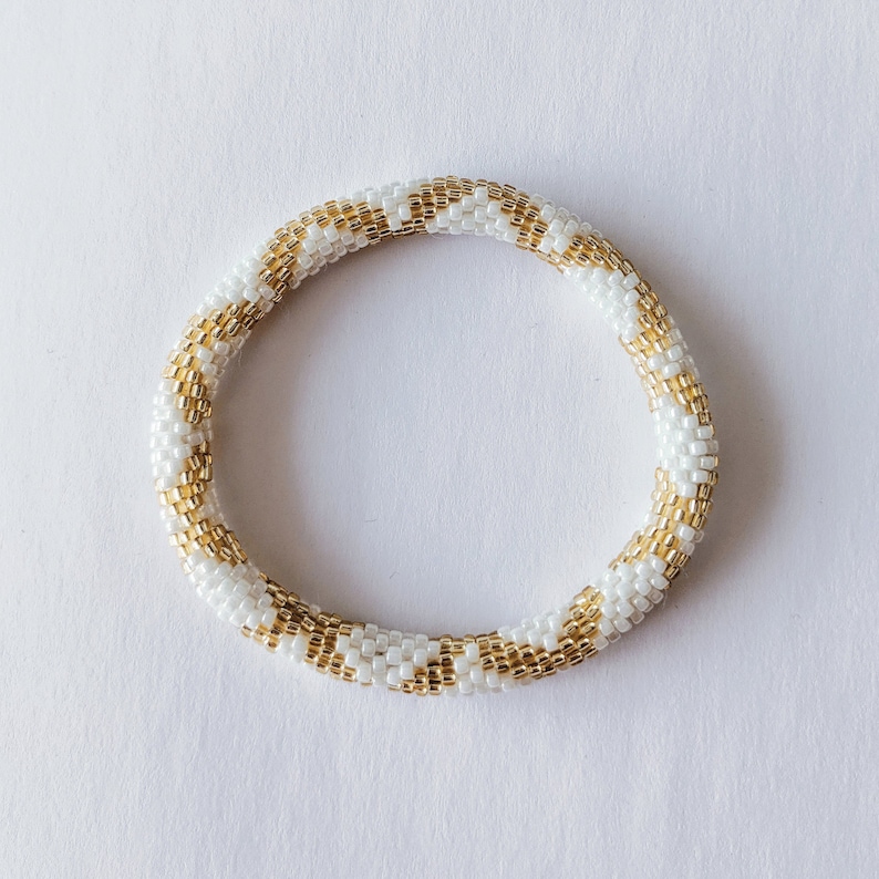 May include: A white and gold beaded bracelet with a chevron pattern.
