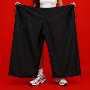 Könnte beinhalten: Übergroße schwarze Hose mit weitem Bein, elastischem Bund mit verstellbaren Kordelzügen und Seitentaschen. Die Hose besteht aus einem leichten, glatten Stoff und verfügt über verstellbare Kordelzüge an den Beinabschlüssen.