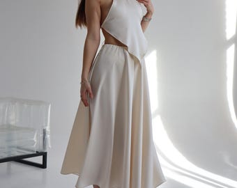 Puede incluir: Conjunto de dos piezas color crema con top halter y falda midi. El top tiene un diseño de espalda abierta. La falda es de estilo fluido y línea A. El conjunto se combina con sandalias de tacón doradas.