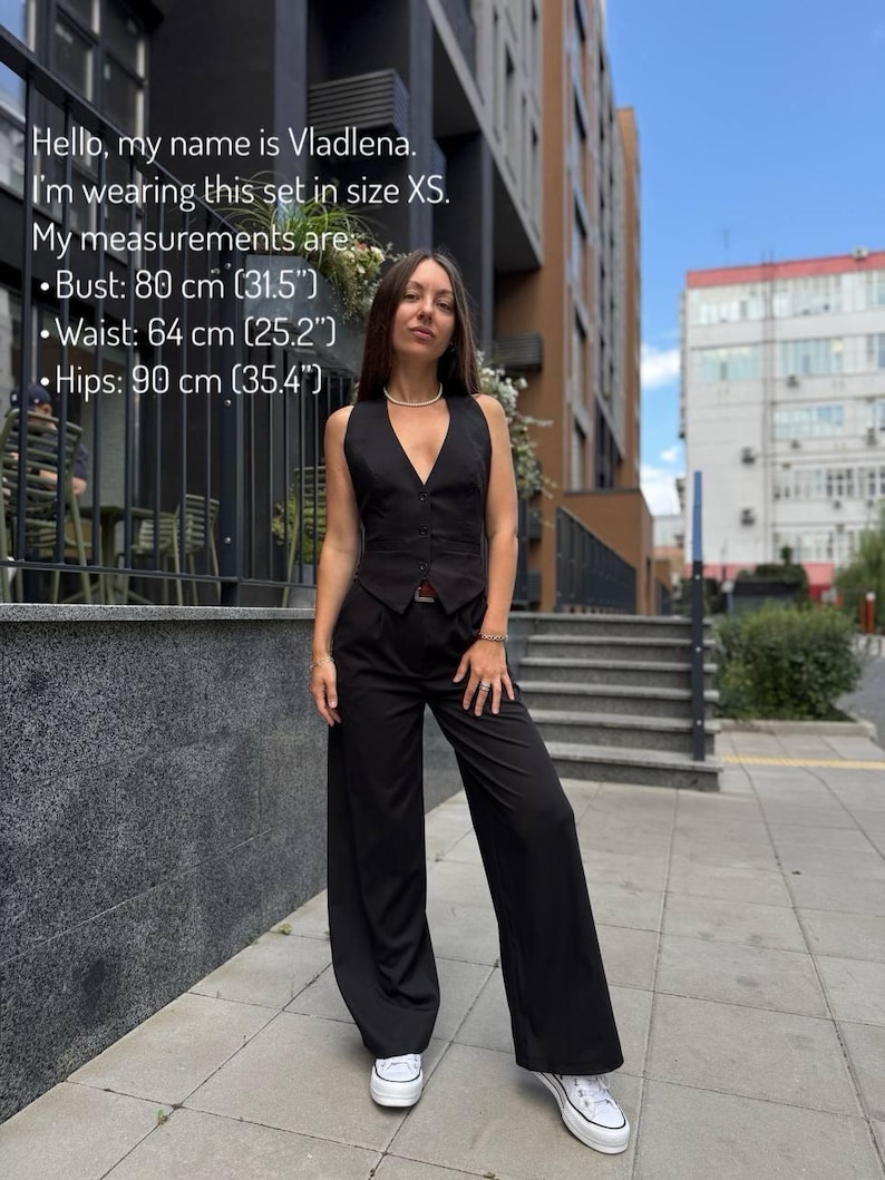 A woman wearing a black jumpsuit is standing on a sidewalk in front of a building.