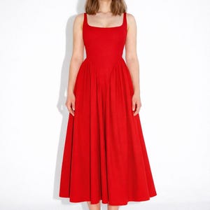 Può includere: Un abito rosso acceso con scollatura quadrata e spalline larghe. L'abito ha un corpetto aderente e una gonna ampia e fluida. La modella indossa sandali con cinturini blu. L'abito è di colore unito e sembra essere realizzato in un tessuto leggero.