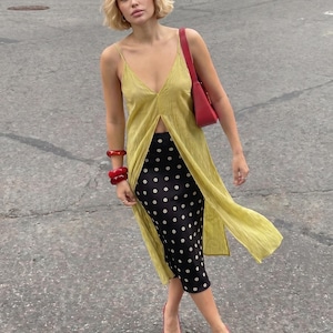 Könnte beinhalten: Eine Frau trägt ein gelbes Kleid mit Schlitz, einen schwarzen Rock mit weißen Punkten und rote Absätze. Sie trägt rote Armbänder und eine rote Handtasche. Das Outfit ist modisch und stilvoll.