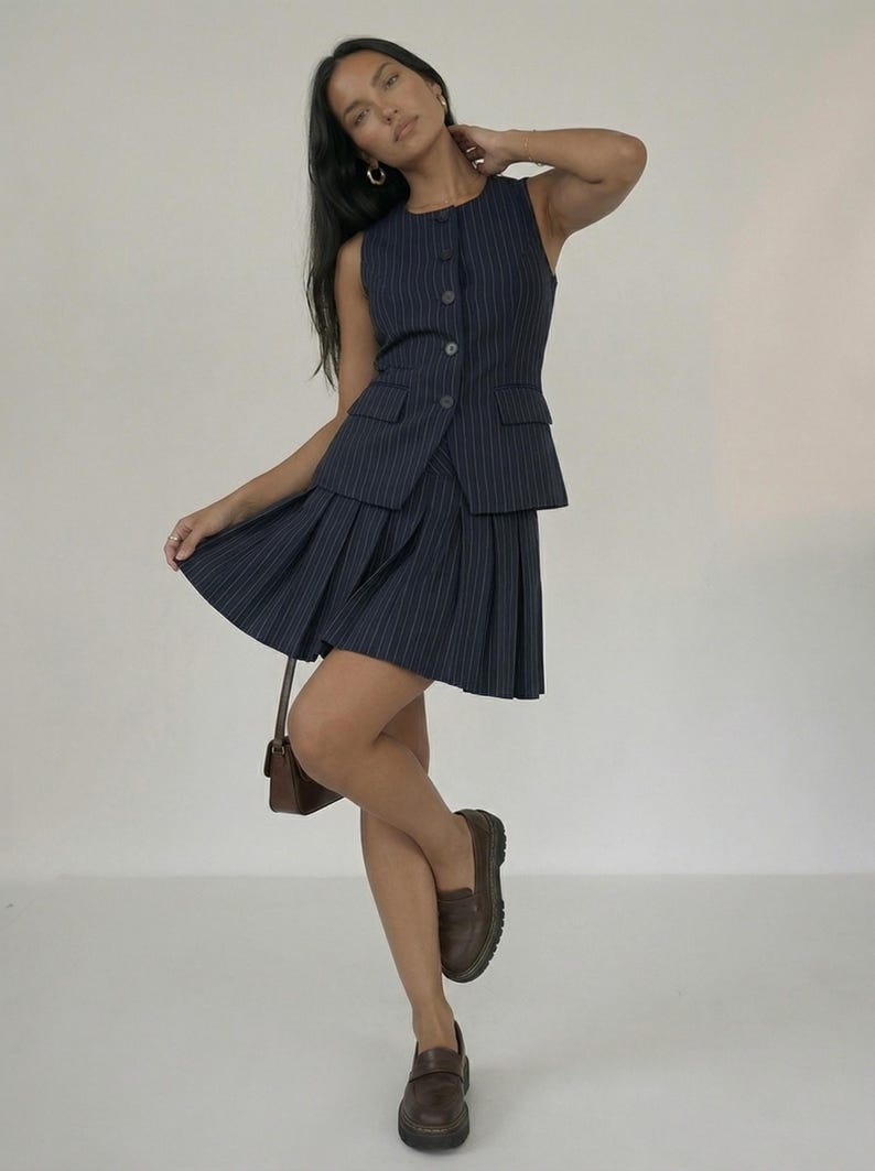 May include: A navy blue pinstriped sleeveless vest with a button-down front and two flap pockets, paired with a matching navy blue pinstriped pleated mini skirt. The outfit is accessorized with a small brown handbag and dark brown loafers.