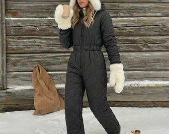 May include: A black quilted jumpsuit with a front zipper, paired with a white faux fur hat and matching white faux fur mittens. The outfit includes tan slip-on shoes, dark sunglasses, and a tan water bottle, creating a winter-ready ensemble.