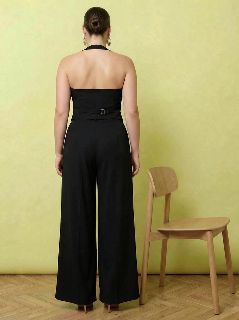 May include: A black halter-style jumpsuit featuring a backless design, a fitted bodice with a waist buckle detail, and wide-leg trousers. The garment is shown from the back, highlighting the open upper back and the clean, tailored silhouette.