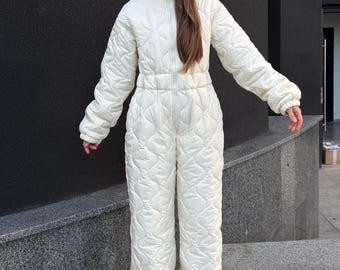 May include: A cream-colored, full-length quilted jumpsuit featuring a front zipper closure, long sleeves with elastic cuffs, and an attached hood. The garment is constructed with a repeating wave-like stitched pattern throughout the entire exterior.