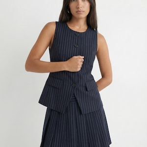 May include: Navy blue pinstripe vest with matching pleated skirt. The sleeveless vest features a button-up front and two small pockets. The skirt has a pleated design. The outfit is a classic business style.
