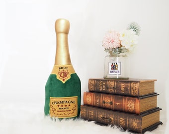 May include: A plush champagne bottle with a green velvet cover and gold accents. The bottle has a label that reads "Champagne Brut France Grande Cuvee". The bottle is sitting on a stack of vintage books with the words "Happy Love" and "Amie" visible on the spines.