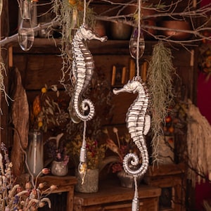 May include: Two silver seahorse-shaped pendant lights hang from a rustic wooden branch with dried flowers and greenery. The lights have clear glass bulbs and are suspended by twine.