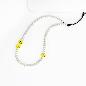 May include: A white beaded phone charm with three yellow smiley face charms. The phone charm is attached to a white device with a black cord.