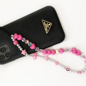 Puede incluir: Un charm de teléfono con cuentas con cuentas de corazón, estrella y carita sonriente rosas. El charm está unido a una funda de teléfono negra con un logotipo de Prada dorado. Las cuentas están ensartadas en un cordón transparente.