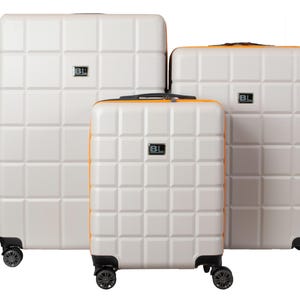 Peut inclure: Ensemble de trois valises rigides blanches avec des accents orange et des roulettes noires. Les valises ont un motif carré et le nom de la marque "BL" est imprimé sur chaque valise.